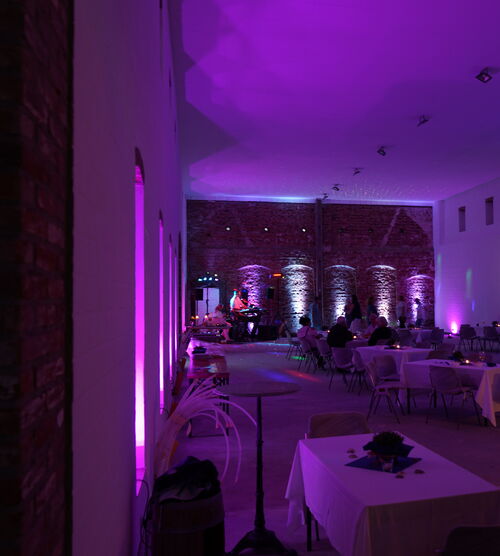Eventscheune bei Nacht. Stühle und viereckige Tische sind aufgebaut. Der Raum wir nur von dekorativen, blauen und violetten LEDs beleuchtet. Einige Personen sitzen an den Tischen.