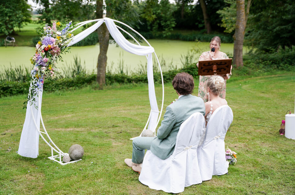 Hochzeitspaar sitzt vor Stoffbespanntem Bogen, Rücken zur Kamrea, schaut nach hinten links. Hintergrund eine Rednerin
