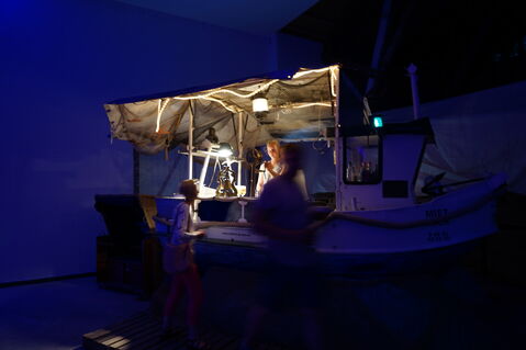 Ein dunkler, leicht blau beleuchteter Raum. Ein Stand in Form eines Fischkutters. Der Stand ist von einer Frau bemannt. Davor stehen zwei verschwommene Personen.