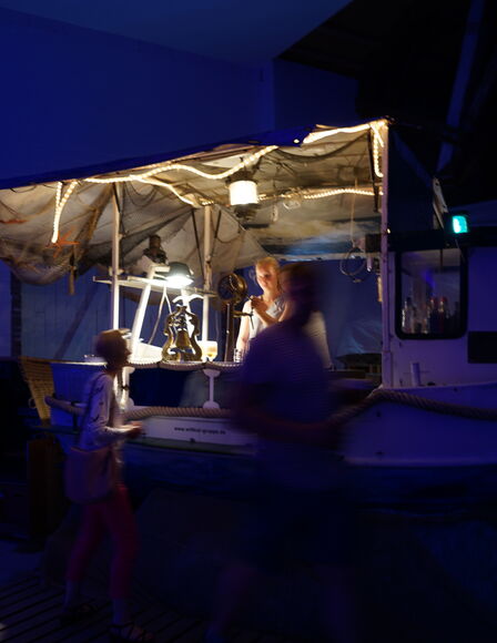 Ein dunkler, leicht blau beleuchteter Raum. Ein Stand in Form eines Fischkutters. Der Stand ist von einer Frau bemannt. Davor stehen zwei verschwommene Personen.